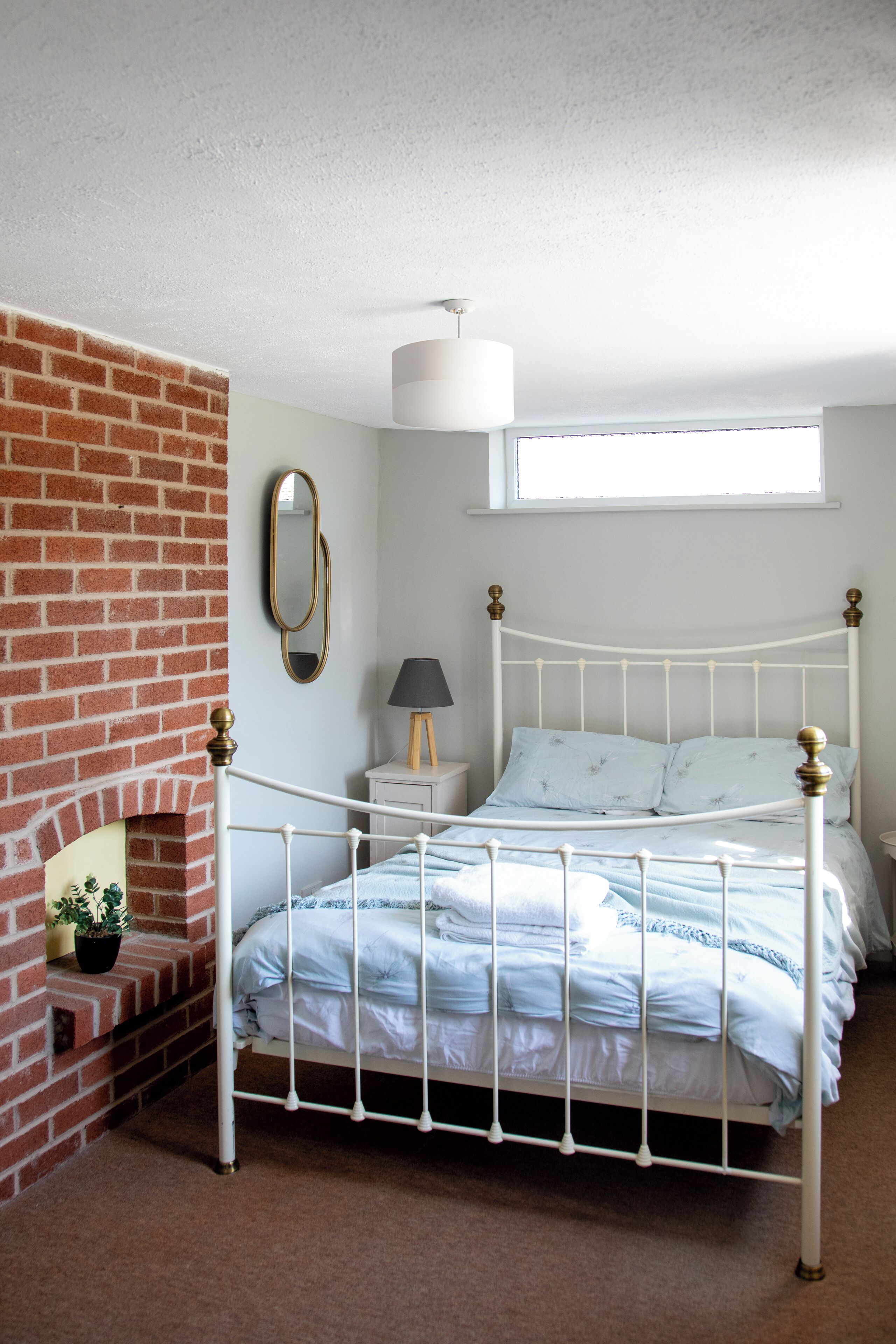 Un dormitorio con estructura de cama metálica blanca, chimenea de ladrillo, mesita de noche pequeña con lámpara y una ventana sobre la cama.