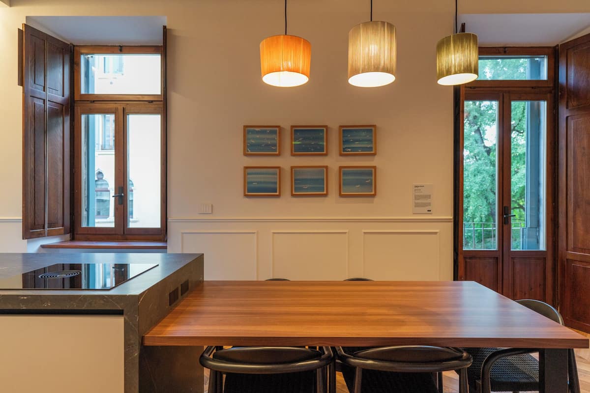 A modern kitchen with a wooden dining table, bar stools, pendant lights, and framed artwork on the wall.