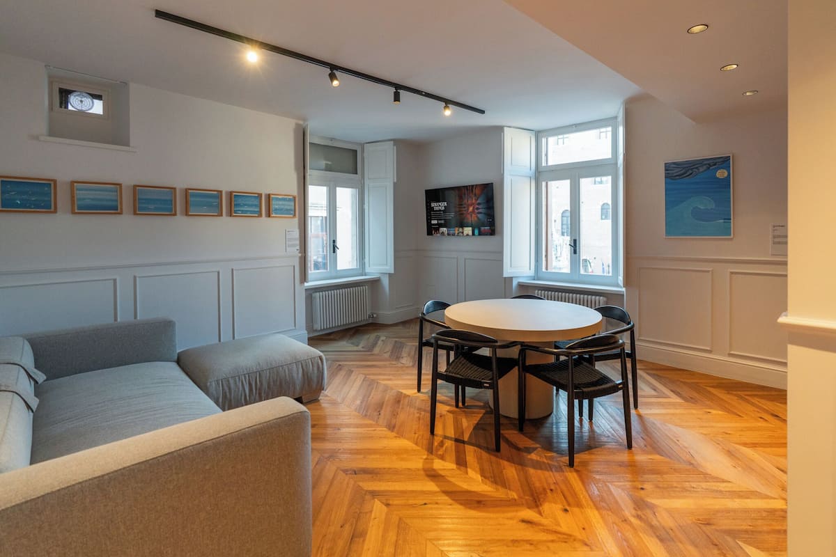 A modern living room with a sofa, a round table with chairs, wooden flooring, and a large window with a view.