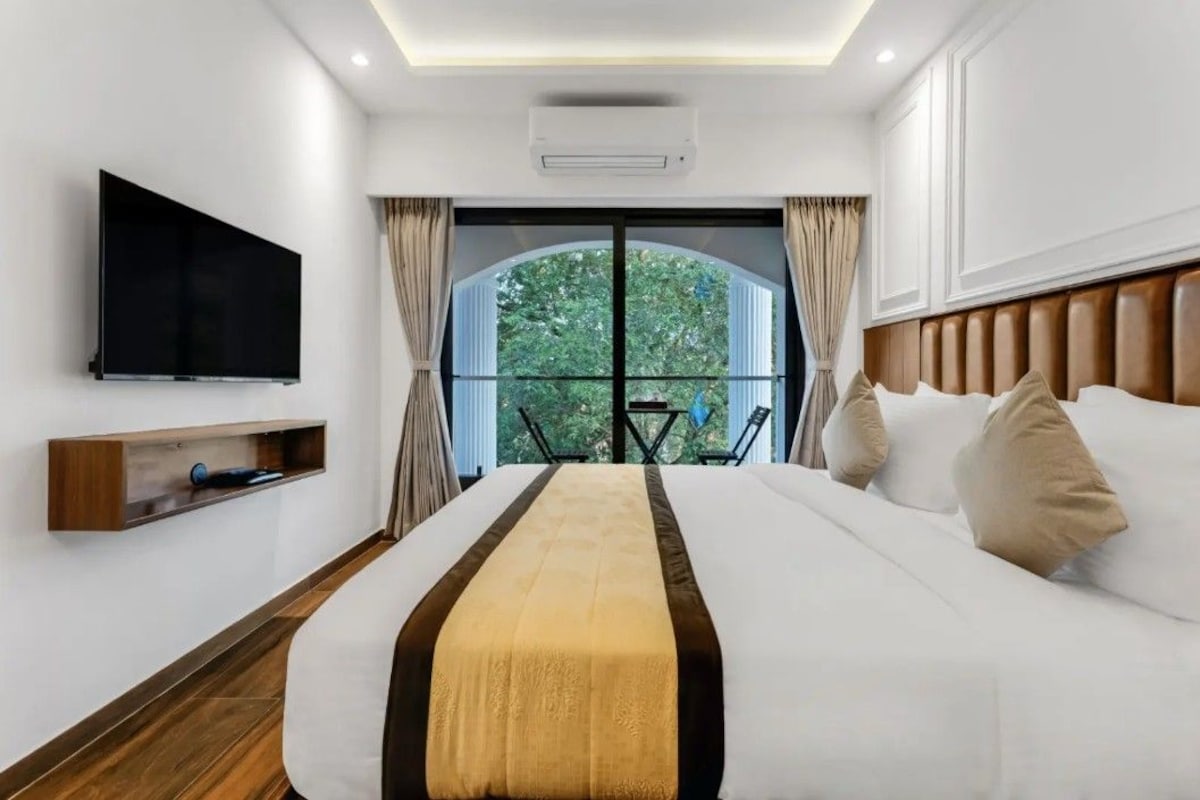 A hotel room with a large bed, a flat-screen TV, a wooden shelf, and a balcony with a view of trees.