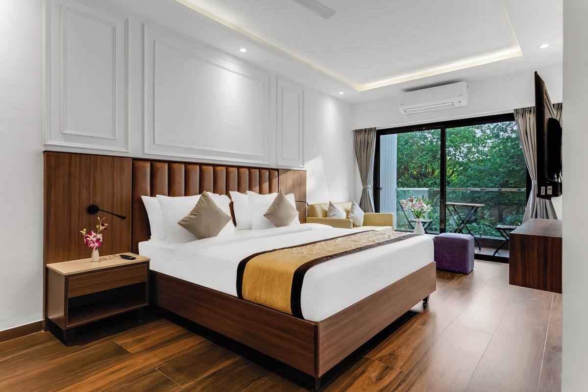 A modern hotel room with a wooden bed, a nightstand, a desk, and a large window offering a view of greenery.