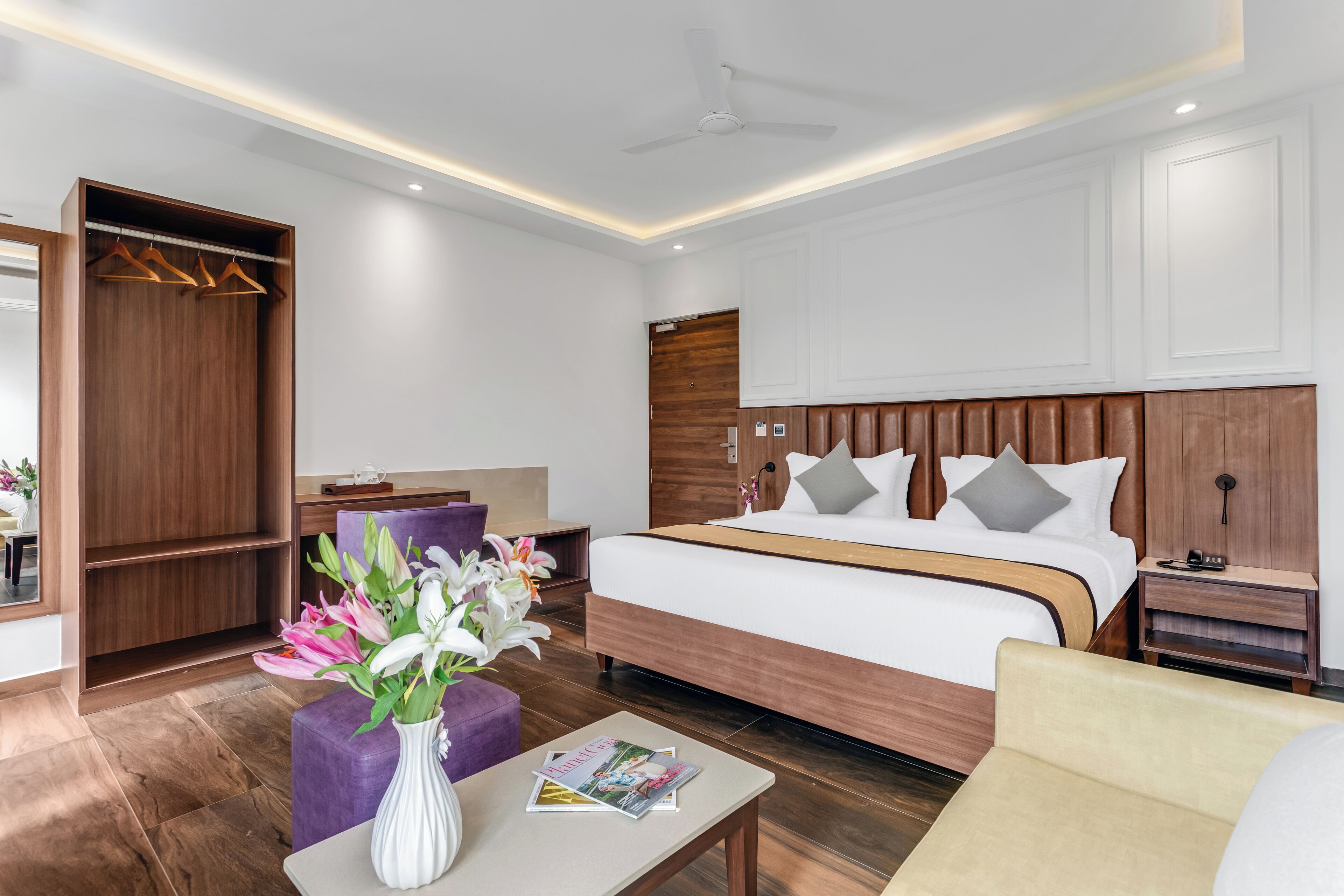 A modern hotel room with a large bed, a wardrobe, a seating area with a table, and a ceiling fan.