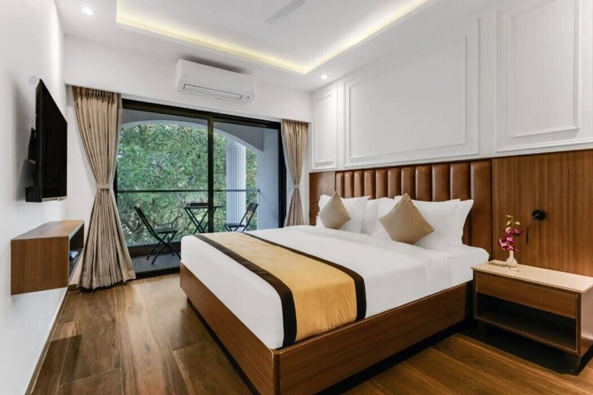 A modern hotel room with a large bed, wooden headboard, a small bedside table, a TV mounted on the wall, and a balcony with a view of greenery.