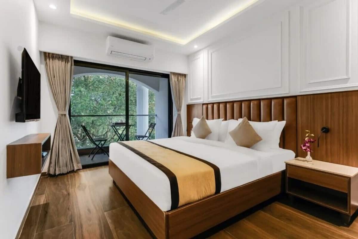A modern hotel room with a large bed, wooden headboard, a small bedside table, a TV mounted on the wall, and a balcony with a view of greenery.