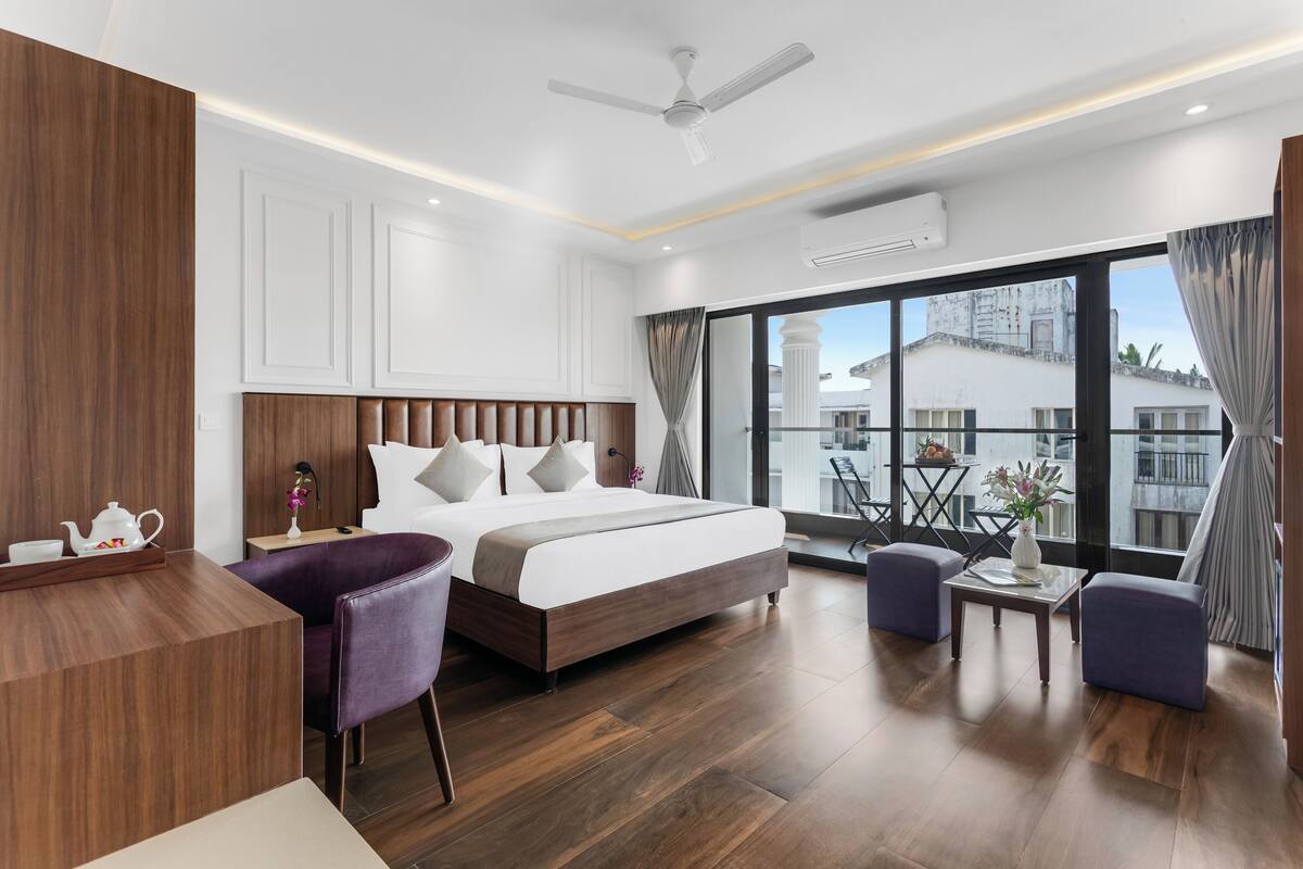 A modern hotel room with a large bed, a desk, a chair, and a balcony with a view of buildings.