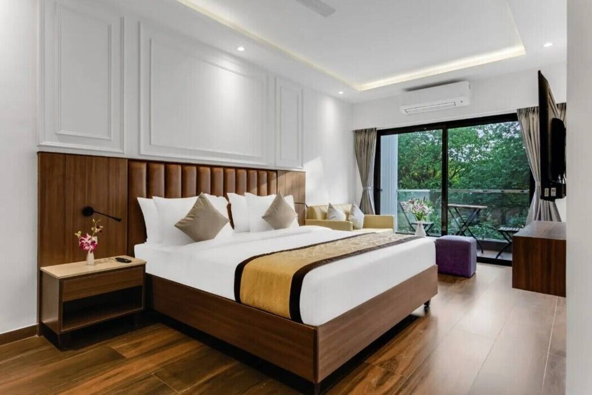 A modern hotel room with a large bed, a nightstand, a TV, and a balcony with a view of greenery.