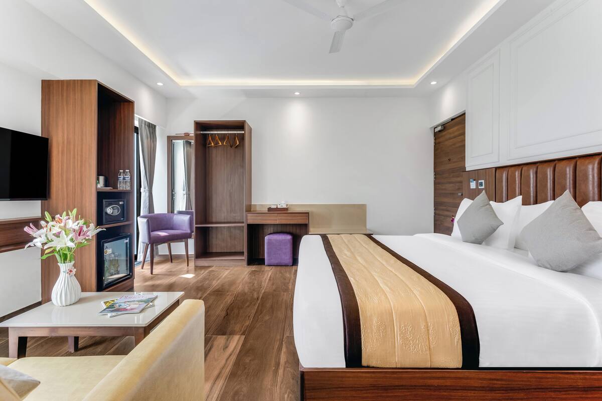 A modern hotel room with a large bed, a desk, a chair, a TV, and a minibar.