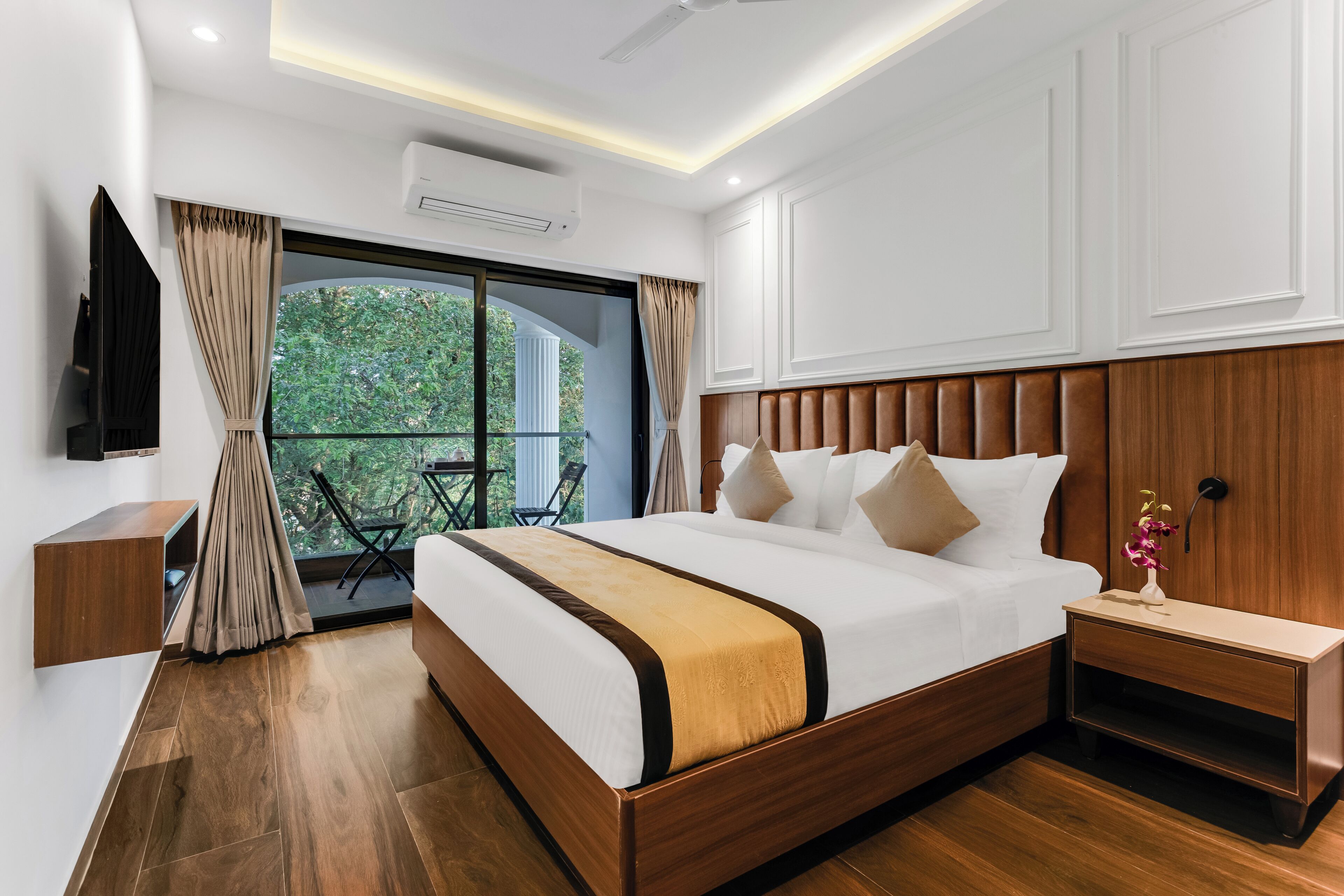 Ein modernes Hotelzimmer mit einem großen Bett, einem Holz-Kopfteil, einem Nachttisch, einem Fernseher und einem Balkon mit Blick ins Grüne.