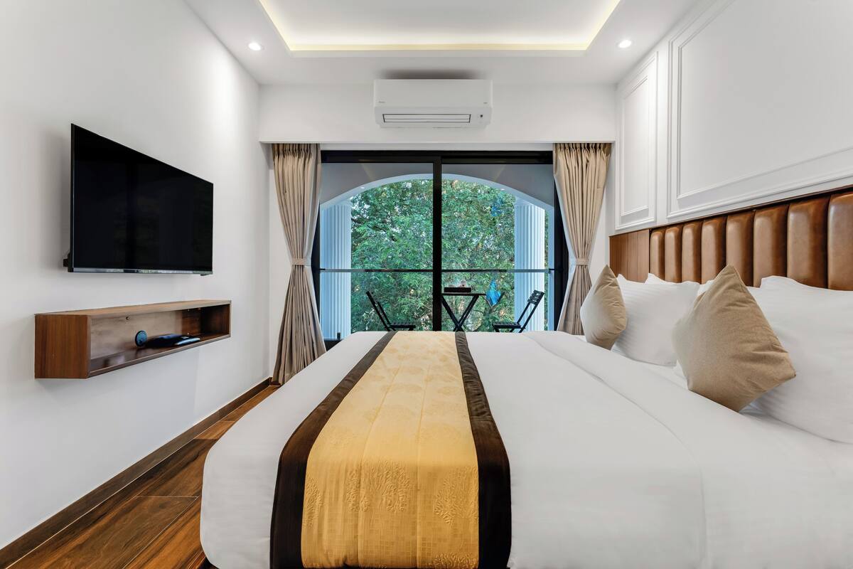 A hotel room with a large bed, a mounted TV, a wooden shelf, and a view of greenery through a glass door.