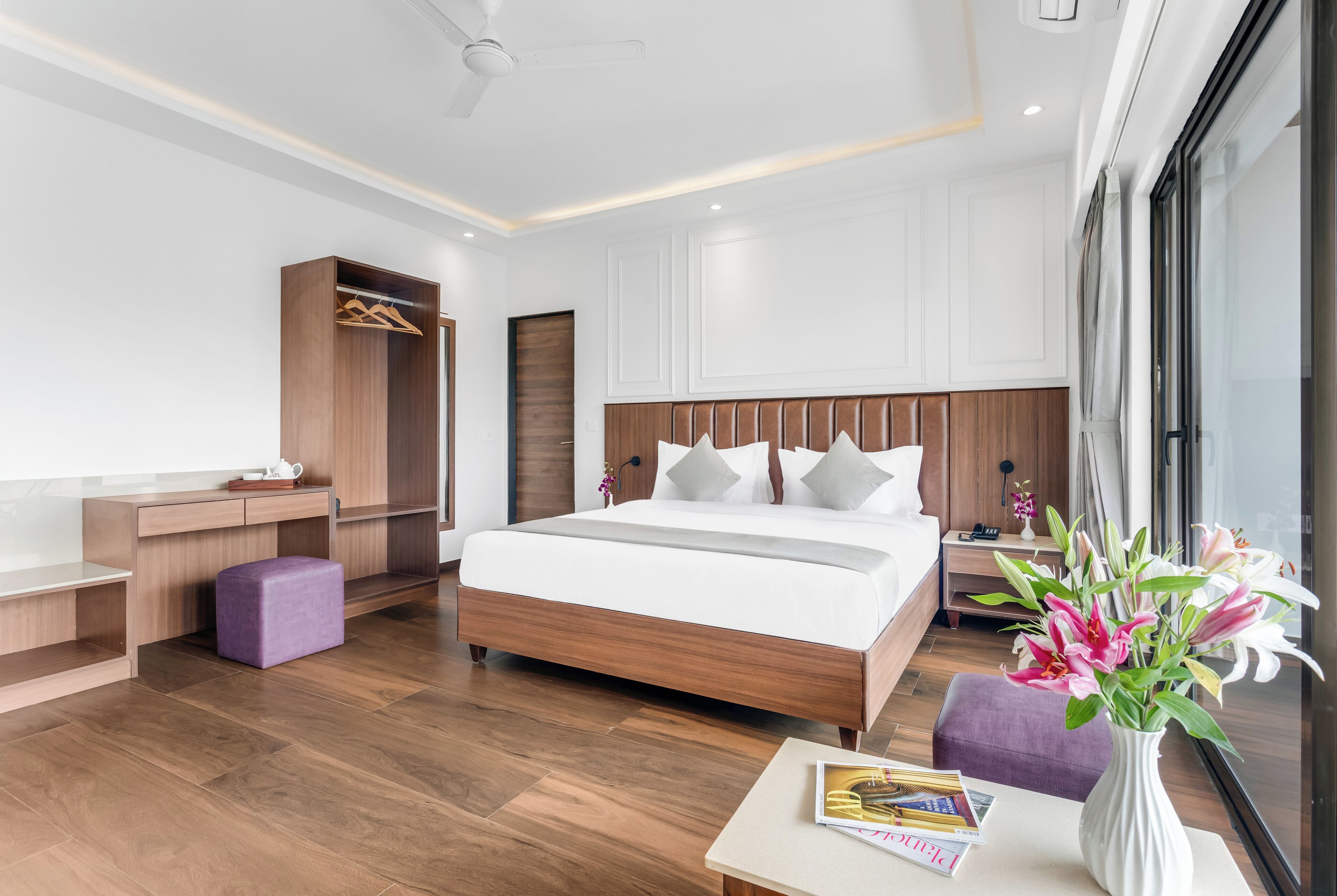 A modern hotel room with a wooden bed, a desk, a wardrobe, and a large window.
