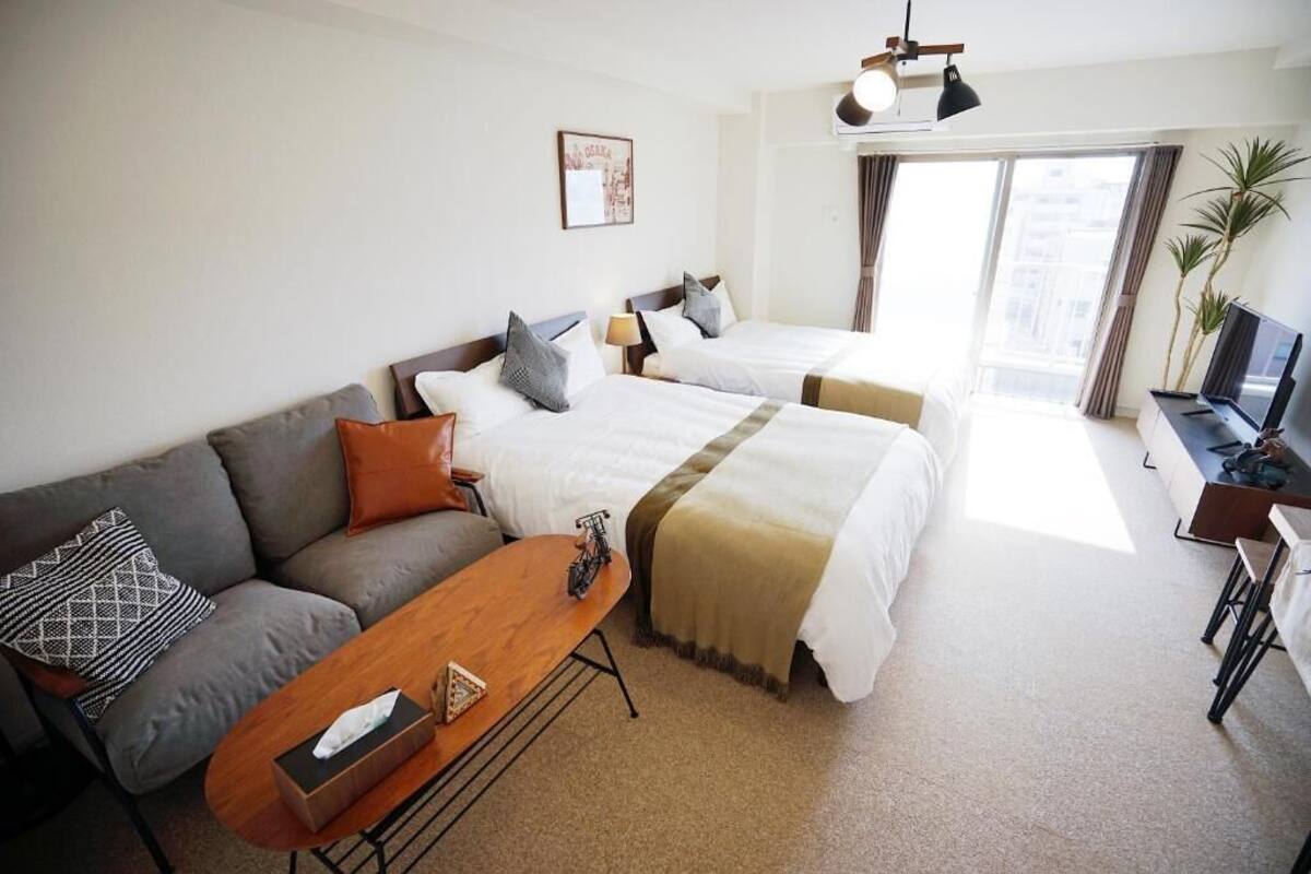 Habitación de hotel con dos camas, un sofá, una mesa de centro, un televisor y un ventanal grande con cortinas.