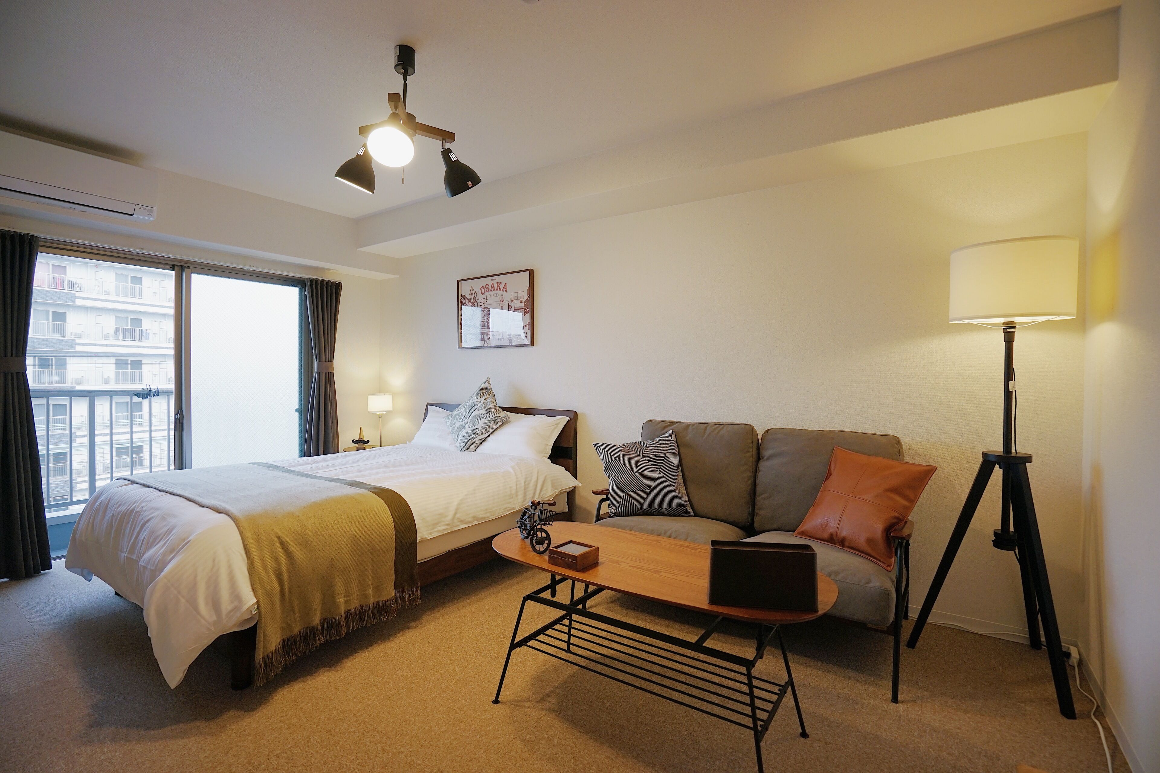 A hotel room with a bed, a sofa, a desk with a laptop, a lamp, and a picture on the wall.