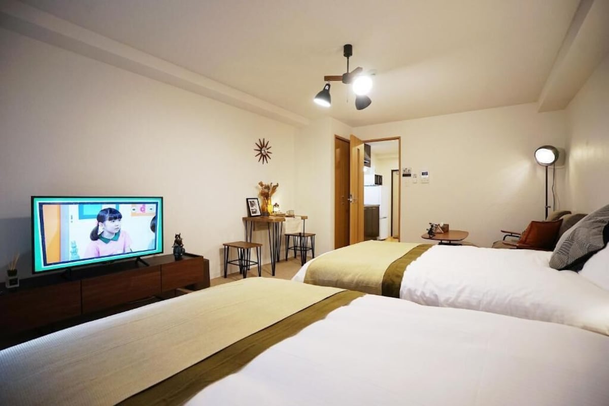Habitación de hotel con dos camas, un escritorio con silla, televisor, ventilador de techo y espejo.