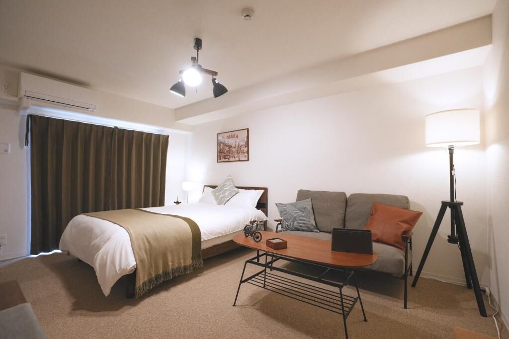 A hotel room with a bed, a sofa, a desk with a laptop, a standing lamp, and a ceiling fan.
