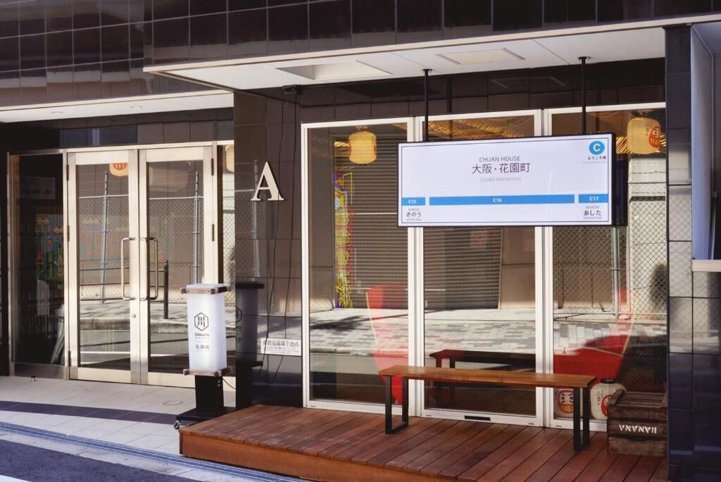 Entrada de hotel con un panel de exhibición digital, un banco de madera en el exterior y un basurero.