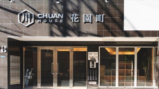Exterior d'un hotel anomenat CHUAN HOUSE amb un disseny modern, que destaca una entrada gran amb portes de vidre i un ascensor visible.