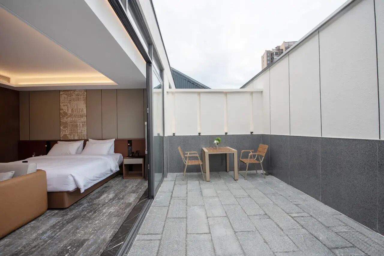 A modern hotel room with a bed, a sofa, a small table with chairs, and a view of the cityscape through large windows.