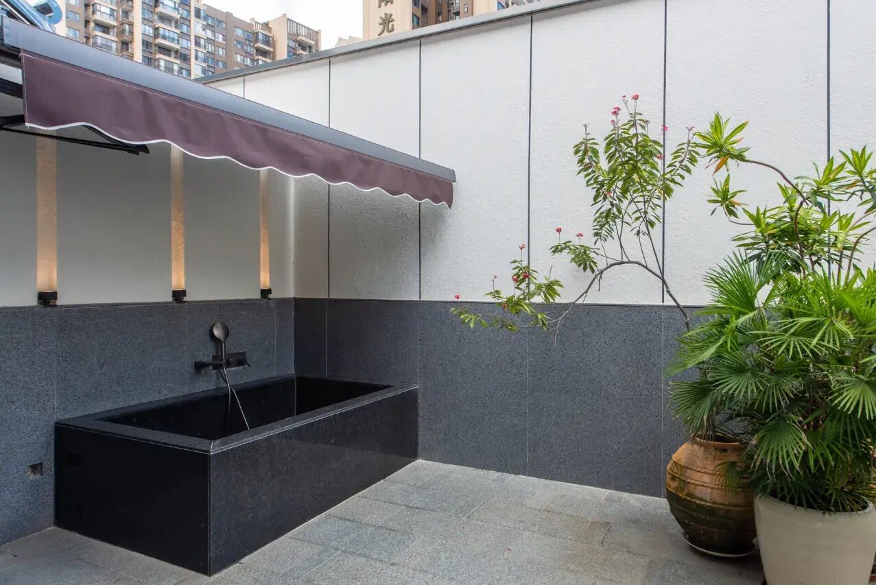 A modern outdoor bathroom with a black bathtub, a decorative awning, and potted plants.