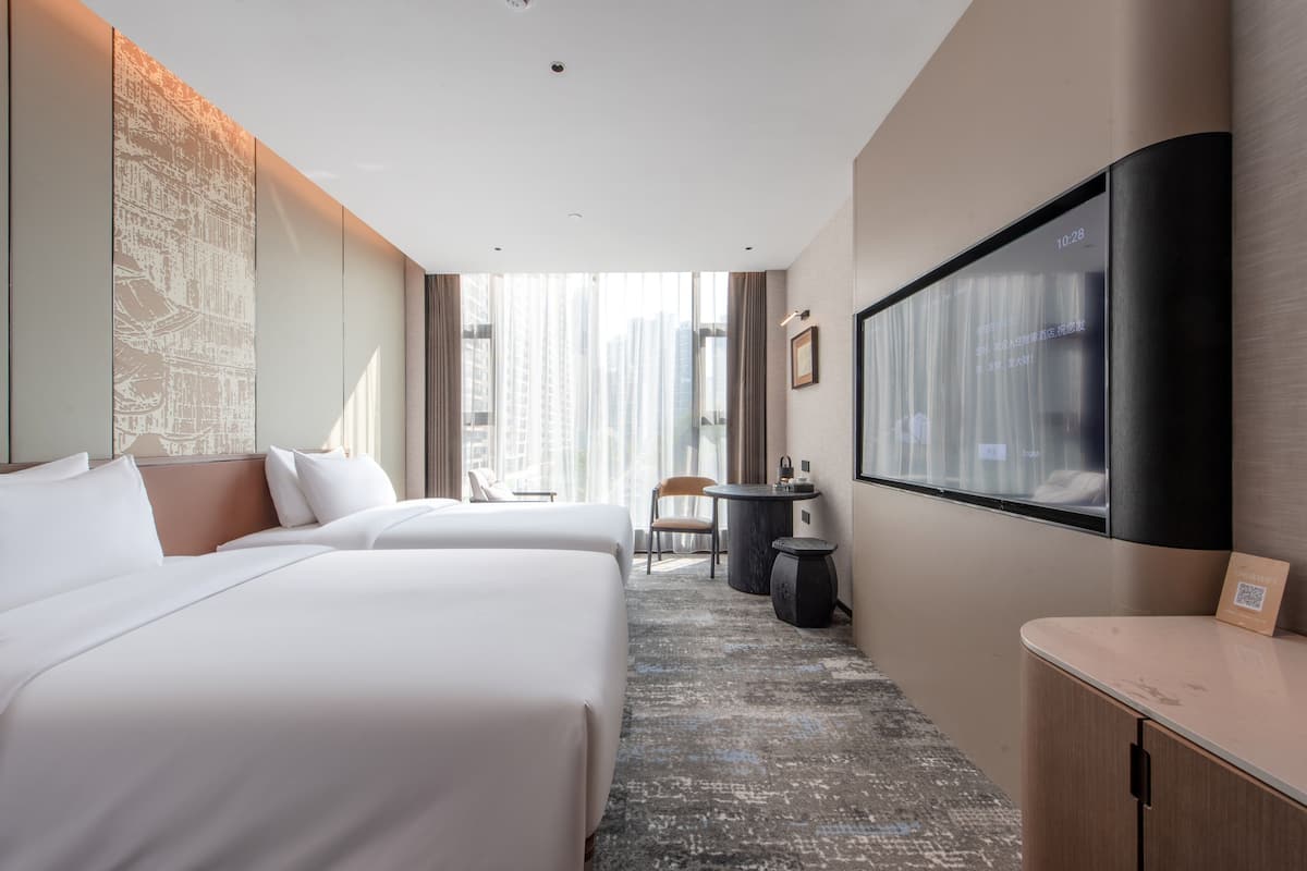 Habitación de hotel con dos camas, un escritorio, una silla, un televisor y un ventanal grande con cortinas.