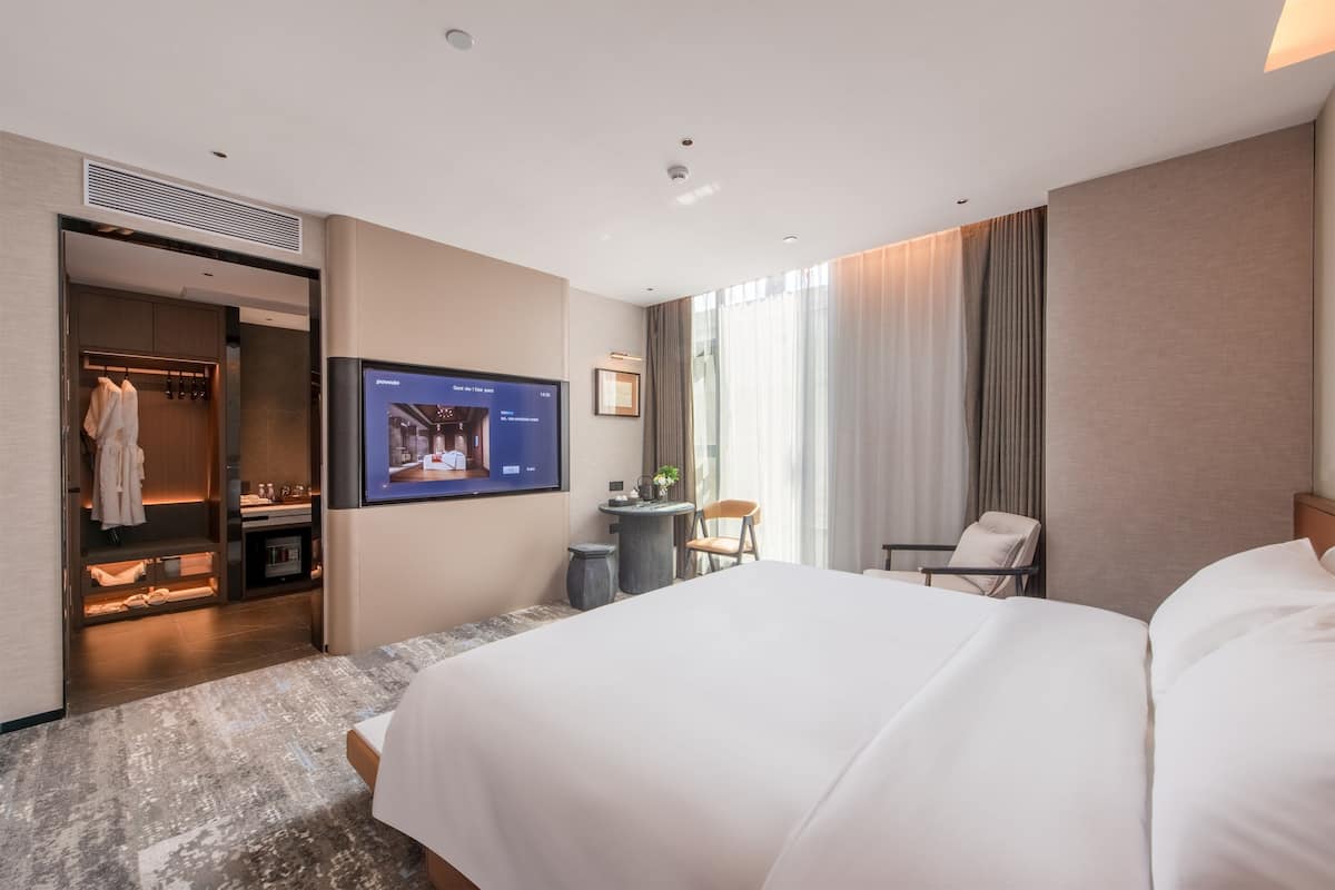 Habitación de hotel con una cama grande, televisor montado en la pared, zona de estar con mesa y sillas, y armario con espejo.