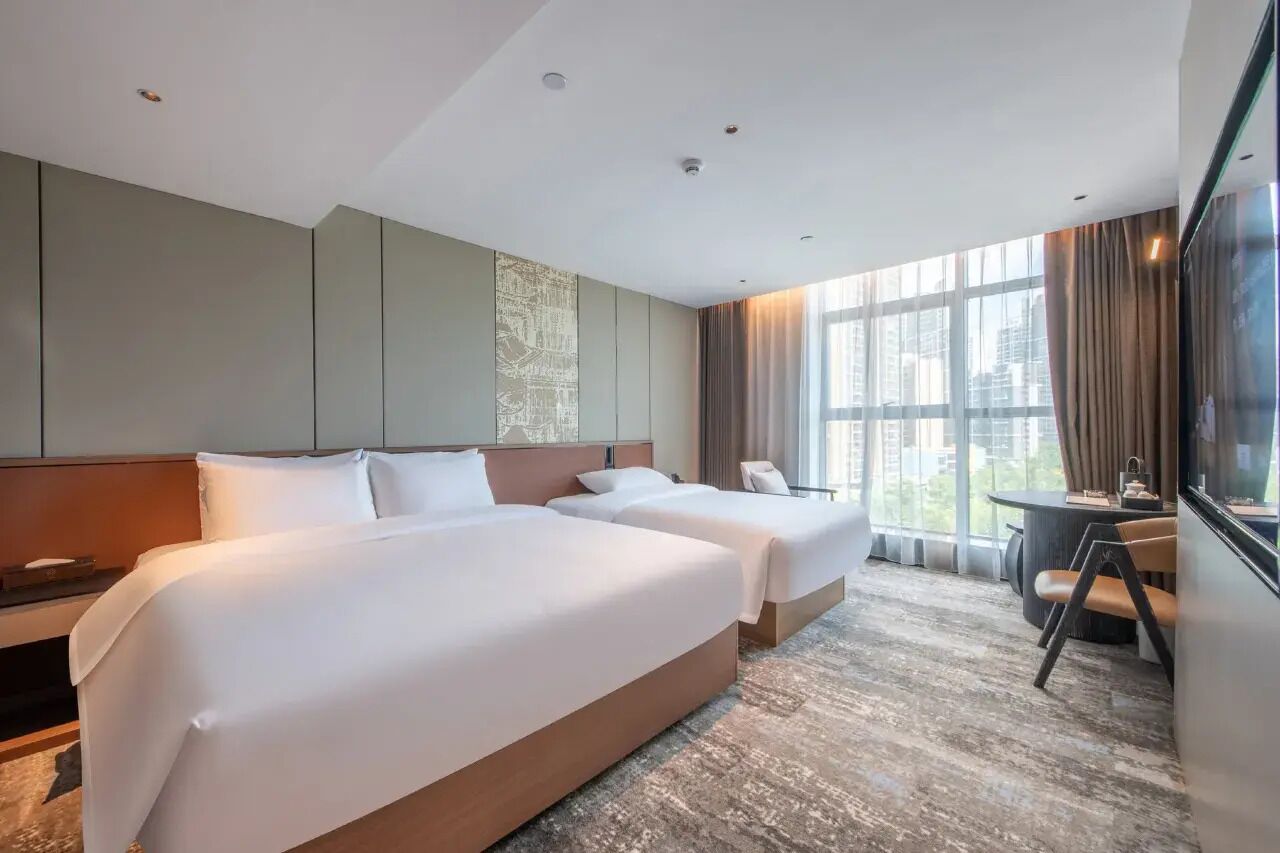 A hotel room with two beds, a desk, and a large window with curtains.
