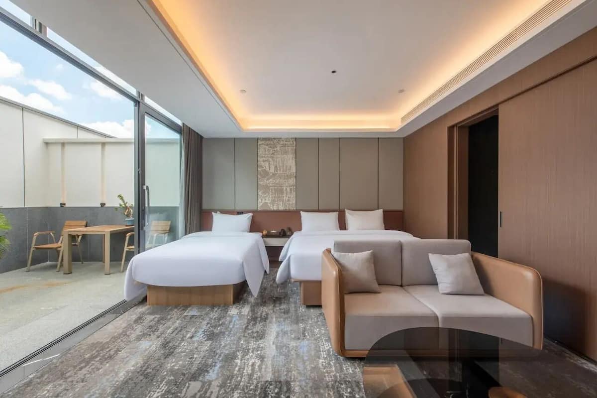 Habitación de hotel moderna con dos camas, un sofá, un comedor con mesa y sillas, y un amplio ventanal con vistas al exterior.