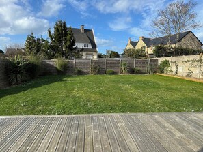 A backyard with a wooden deck, a stone wall, and a grassy lawn. - VILLA ADTHEO (Dinard)