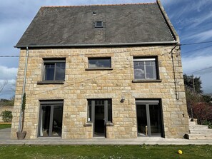 Ein zweistöckiges Steinhaus mit dunklem Dach, großen Fenstern und einer Haustür.