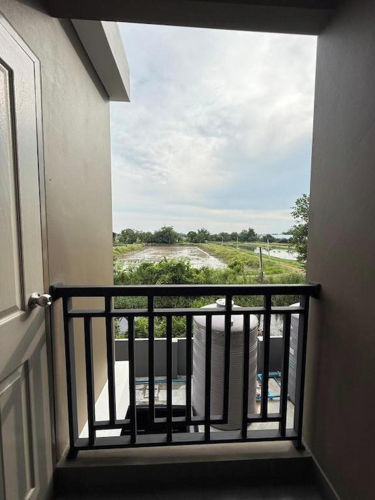 A balcony with a metal railing, a door, and a view of a green landscape with trees and a cloudy sky.