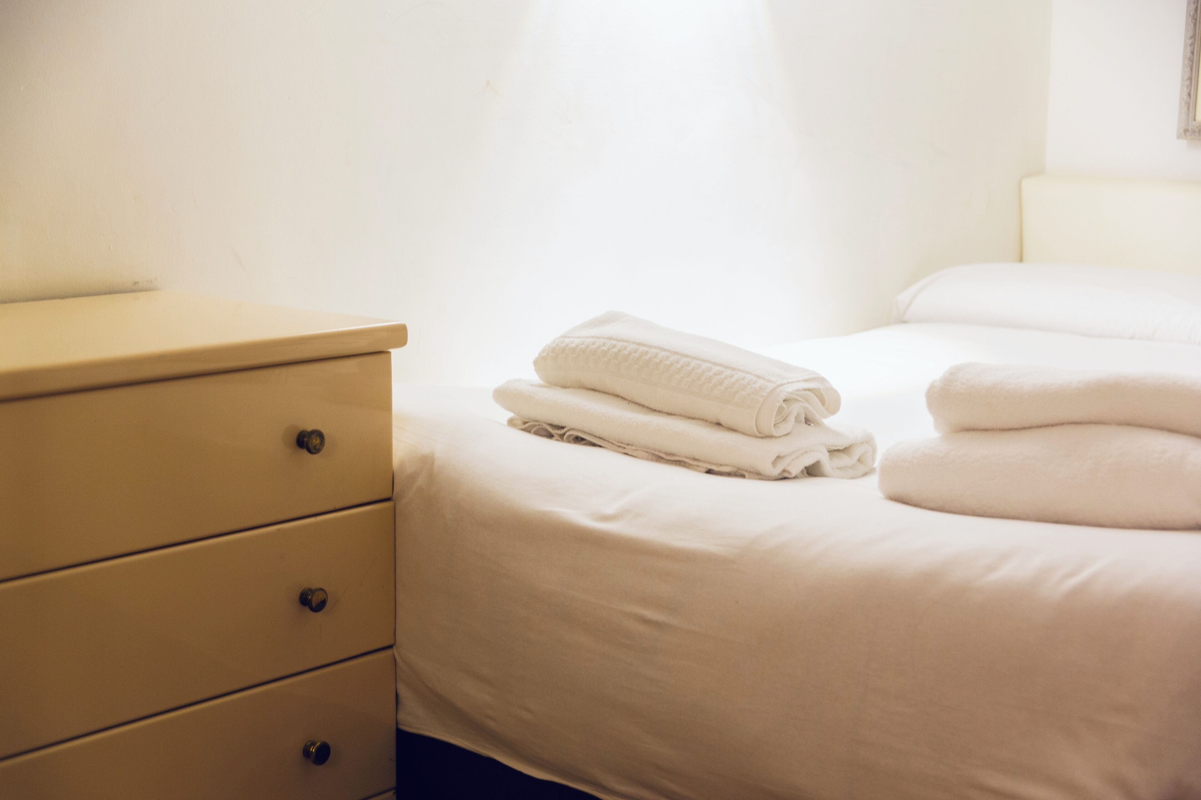 A neatly made bed with folded white linens, a bedside table with drawers, and a plain wall.