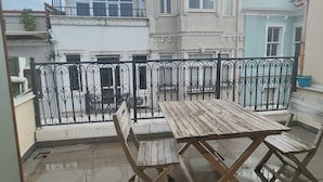 Une cour avec une table et des chaises en bois, une clôture en métal et, au fond, un bâtiment doté de nombreuses fenêtres et de balcons.