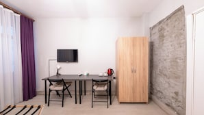 Une petite pièce moderne, dotée d’une armoire en bois, d’un bureau avec deux chaises, d’une télévision et d’une fenêtre ornée de rideaux violets.