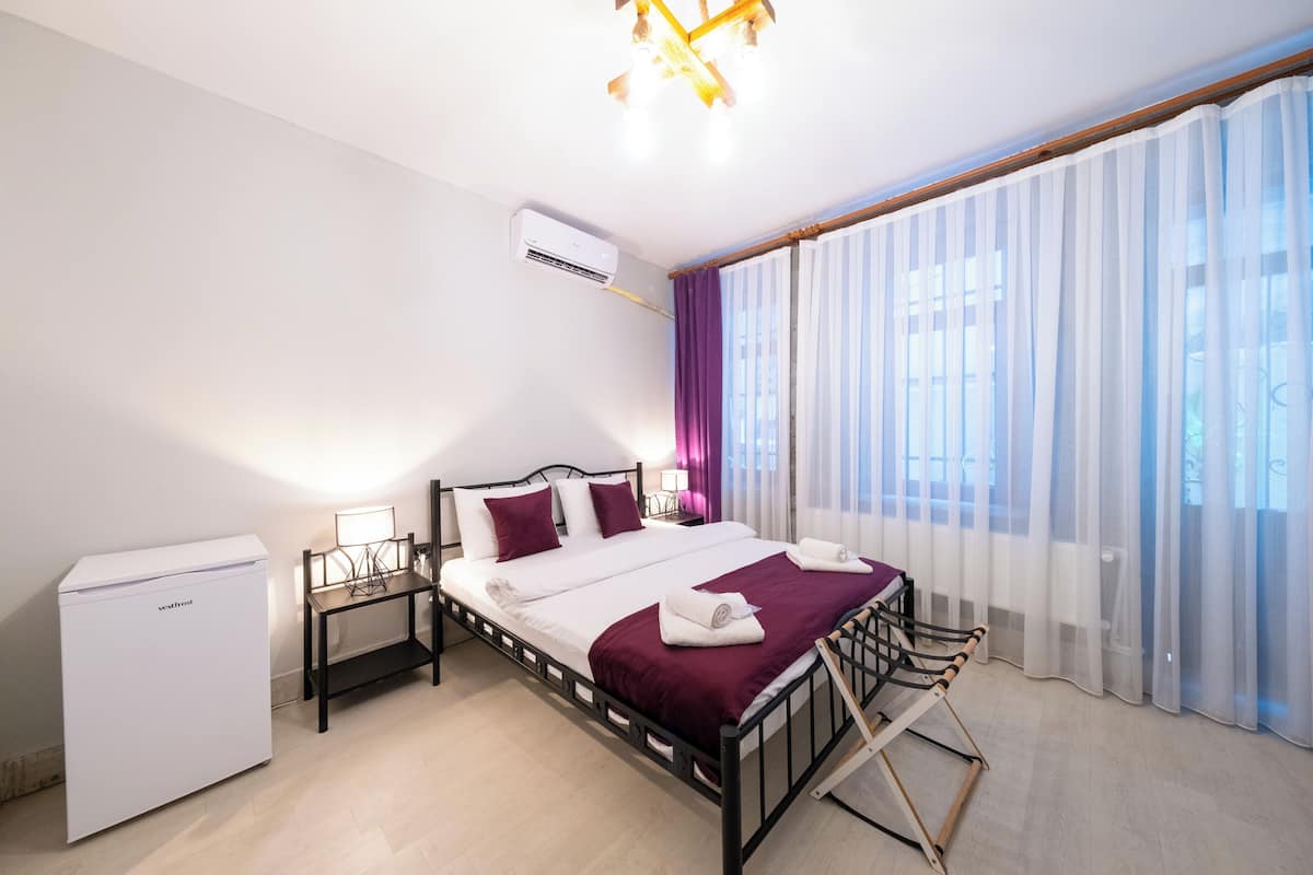 Un dormitorio con cama, mesitas de noche, escritorio, silla, armario y ventana con cortinas.