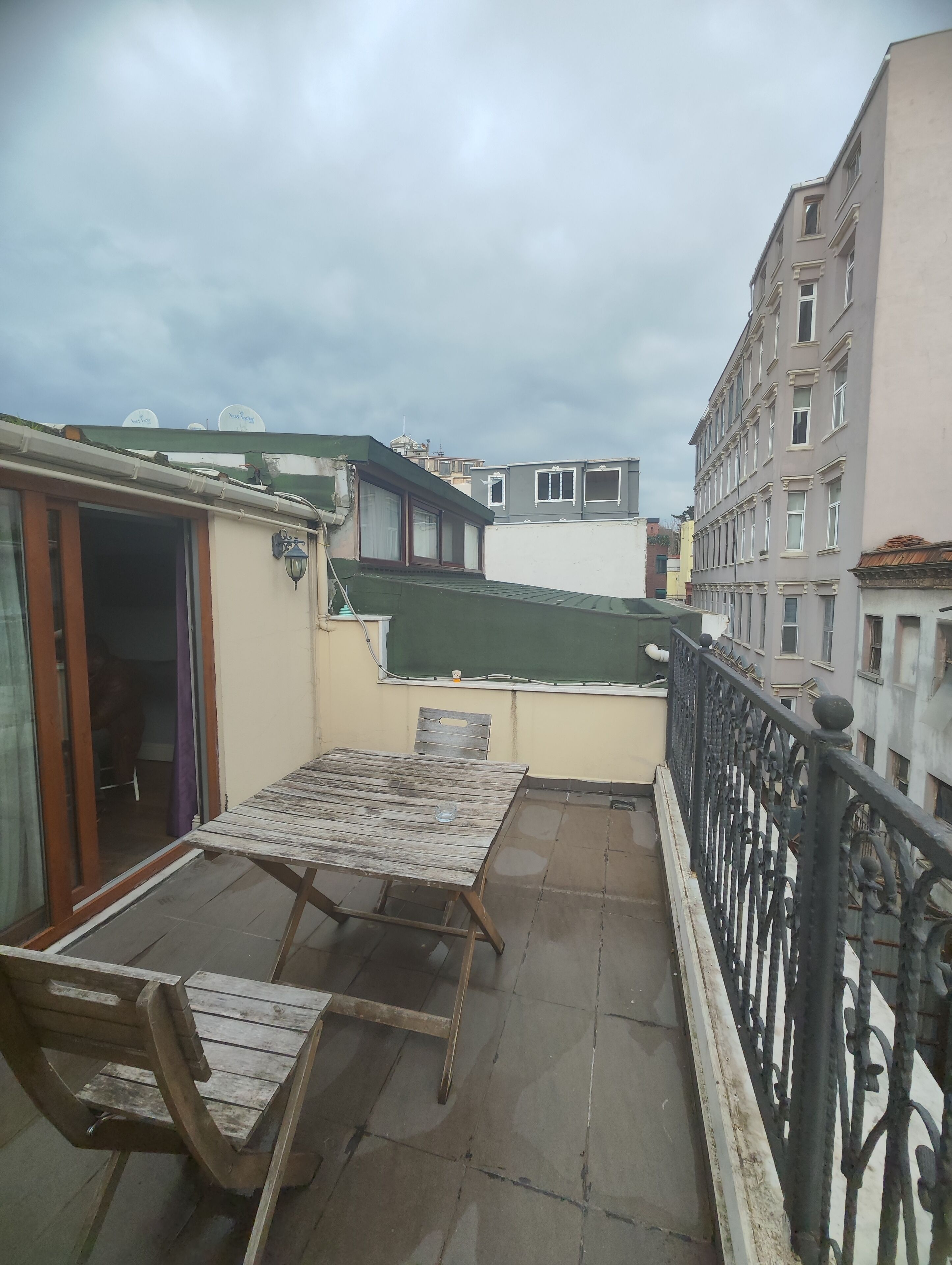 A rooftop terrace with a wooden table and chairs, a green awning, and a view of surrounding buildings.