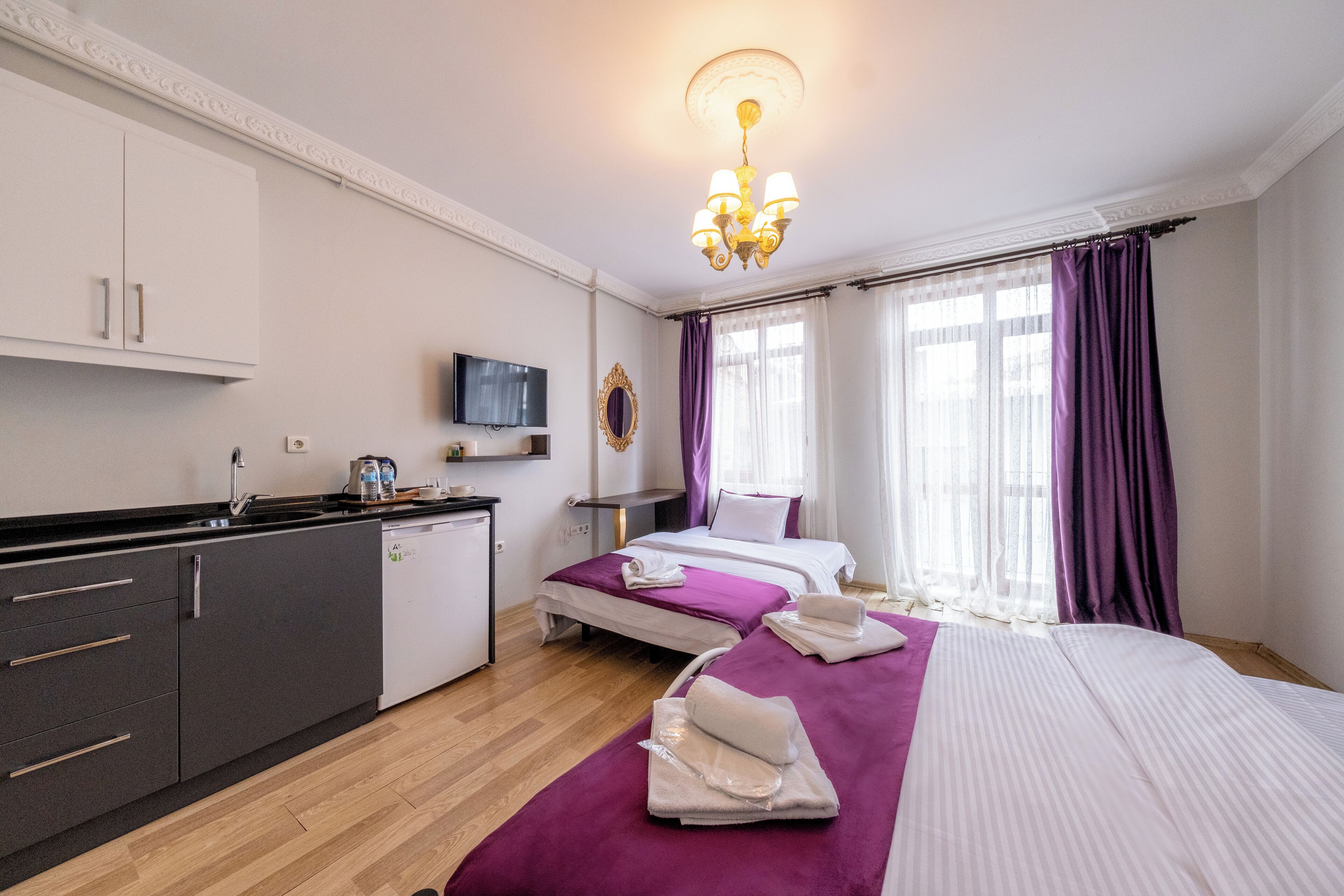 A hotel room with a kitchenette, two beds with purple bedding, a TV, and a chandelier.