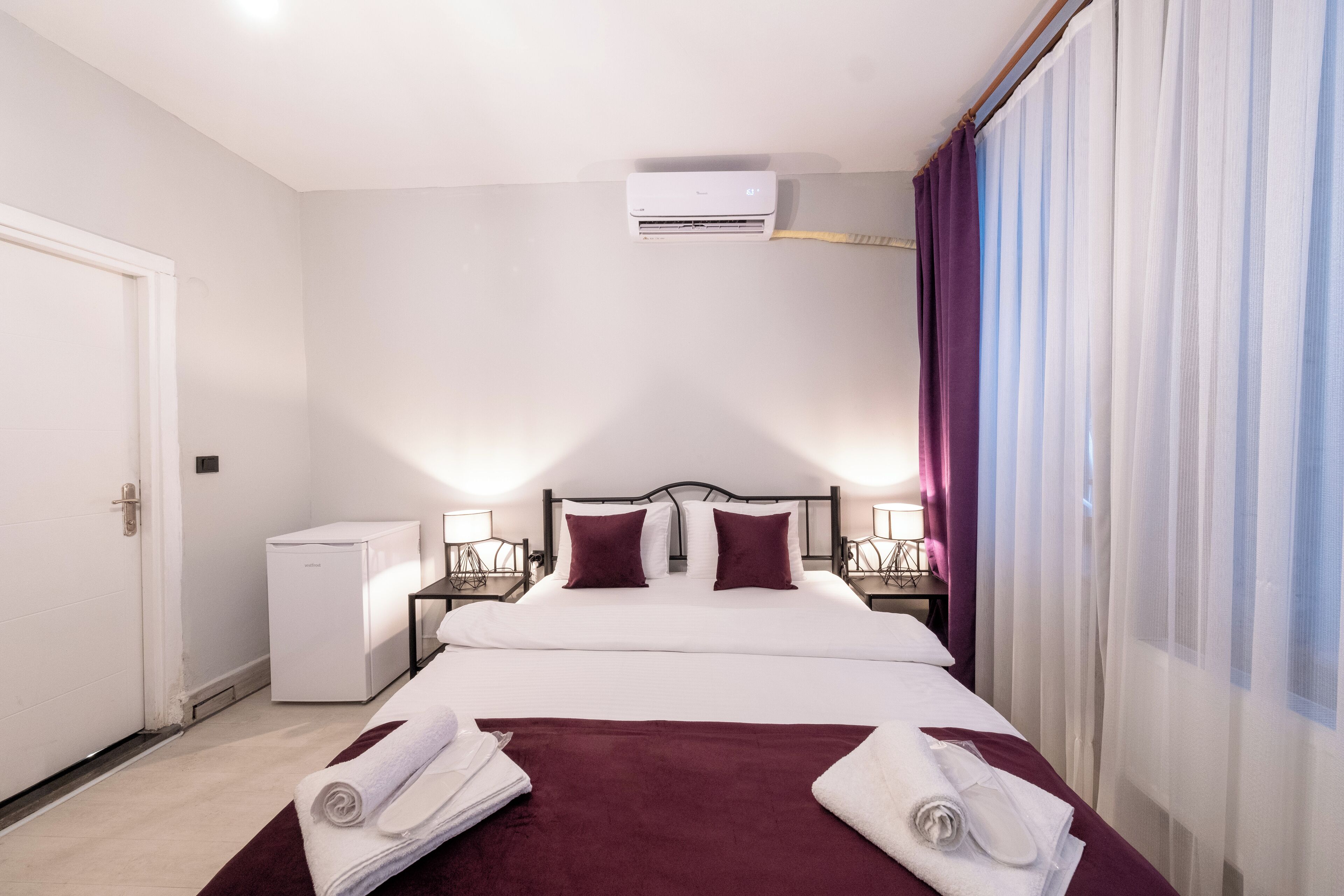 Habitación de hotel con cama, paredes blancas, un refrigerador pequeño blanco y una ventana con cortinas translúcidas.