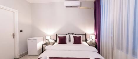 Habitación de hotel con cama, paredes blancas, un refrigerador pequeño blanco y una ventana con cortinas translúcidas.