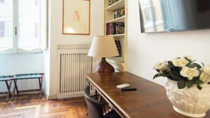 Ein Zimmer mit einem Holztisch, einer Lampe, einer weißen Vase mit Blumen, einem Bücherregal, einem gerahmten Bild und einem Fernseher an der Wand.