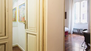 Ein Flur mit einer Tür zu einem Zimmer, das ein Bett, einen Tisch und ein Fenster mit Vorhängen bietet.