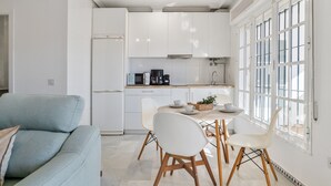 Una cocina moderna con armarios blancos, un comedor con mesa y sillas, y un amplio ventanal con persianas.