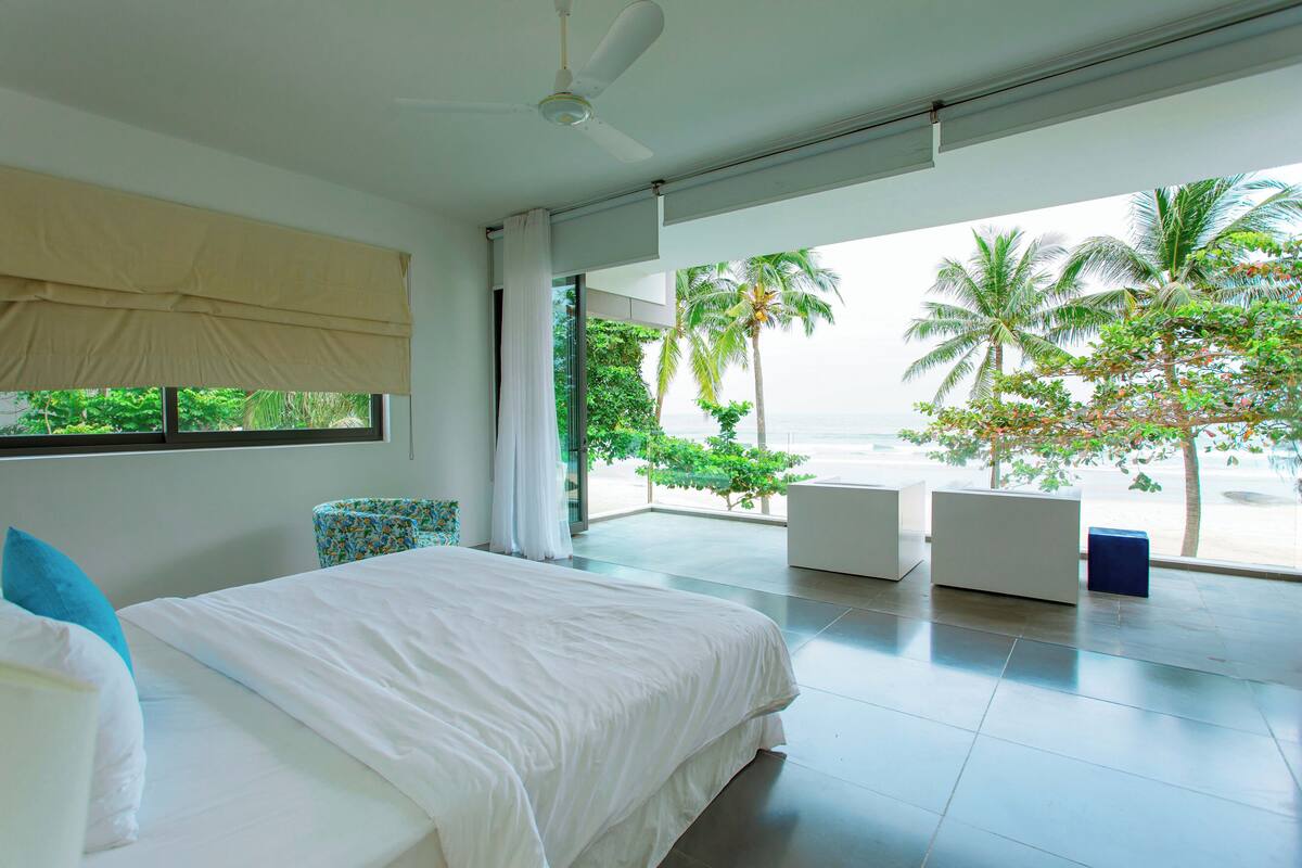 Un dormitorio moderno con una cama grande, un ventilador de techo y vistas al océano y palmeras.