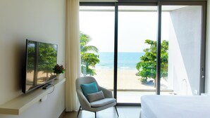Camera d'albergo con un letto, una TV su un mobile, una sedia con cuscino e un'ampia porta-finestra scorrevole con vista sulla spiaggia.