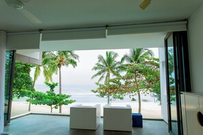 A room with a large sliding glass door leading to a beach view, featuring palm trees and a sandy shore. - Sanctuary Ho Tram Resort – Private Pool Villas (Ho Chi Minh City)