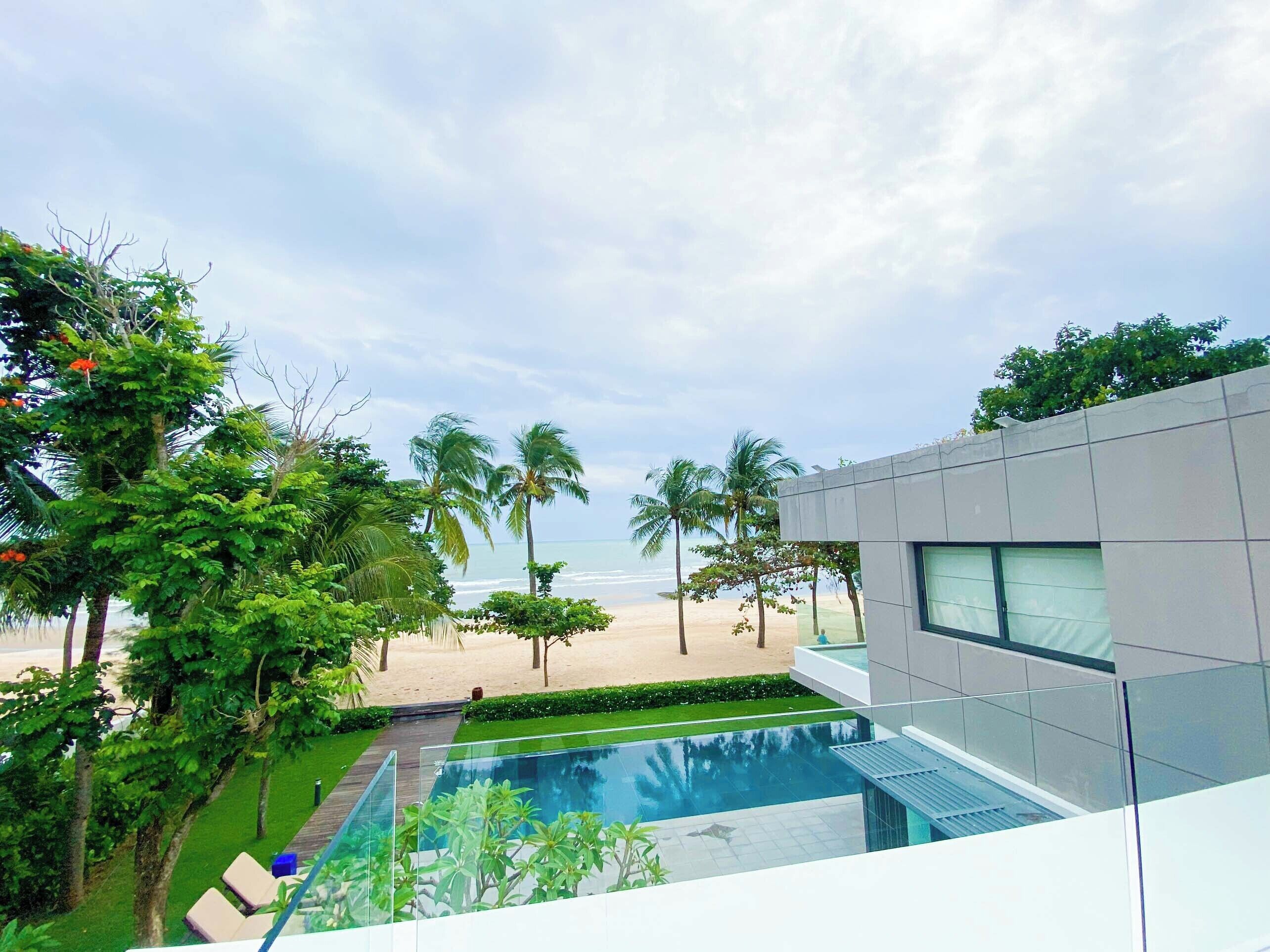 A modern building with a glass pool, palm trees, and a beach view.