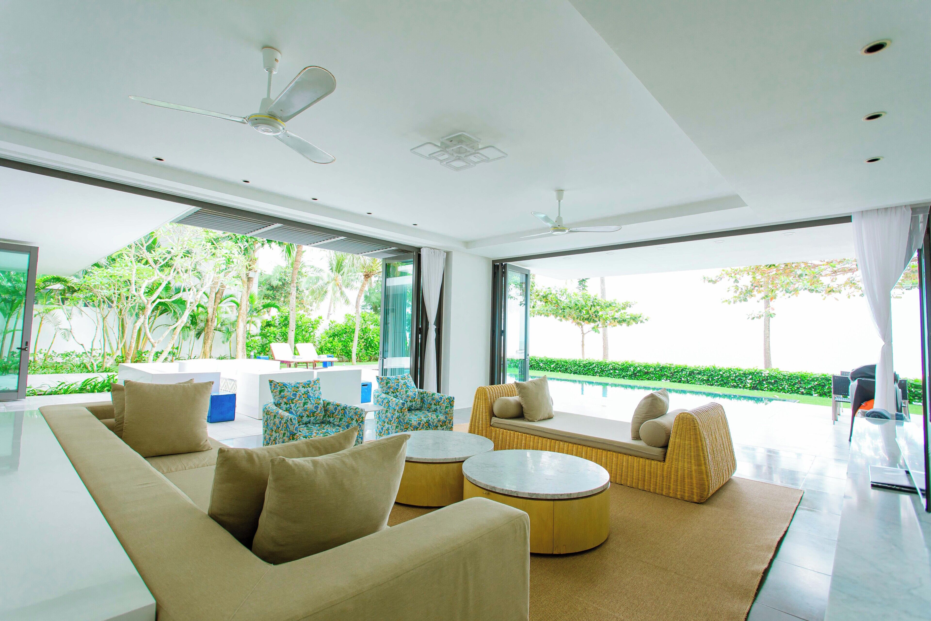 A modern living room with a sofa, armchairs, and a round coffee table. There are ceiling fans and large windows with a view of greenery outside.