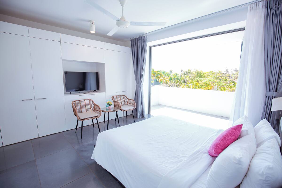 Una habitación de hotel moderna con una cama grande, televisor, dos sillones y un balcón con vistas.