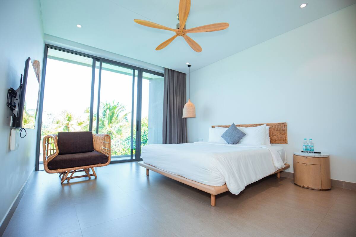 Habitación de hotel moderna con una cama grande, una silla de mimbre, un ventilador de techo y vistas a palmeras a través de la ventana.