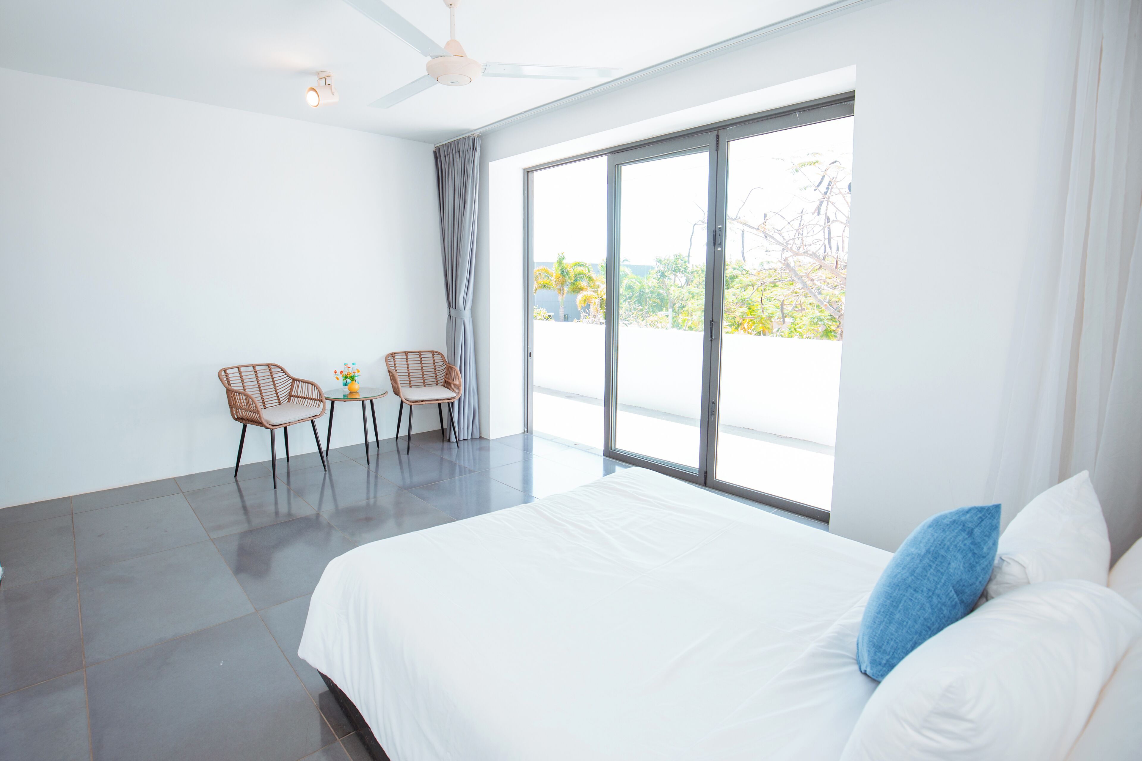 A modern hotel room with a large bed, two chairs, a small table, and a view through large windows.