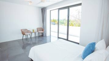 A modern hotel room with a large bed, two chairs, a small table, and a view through large windows.