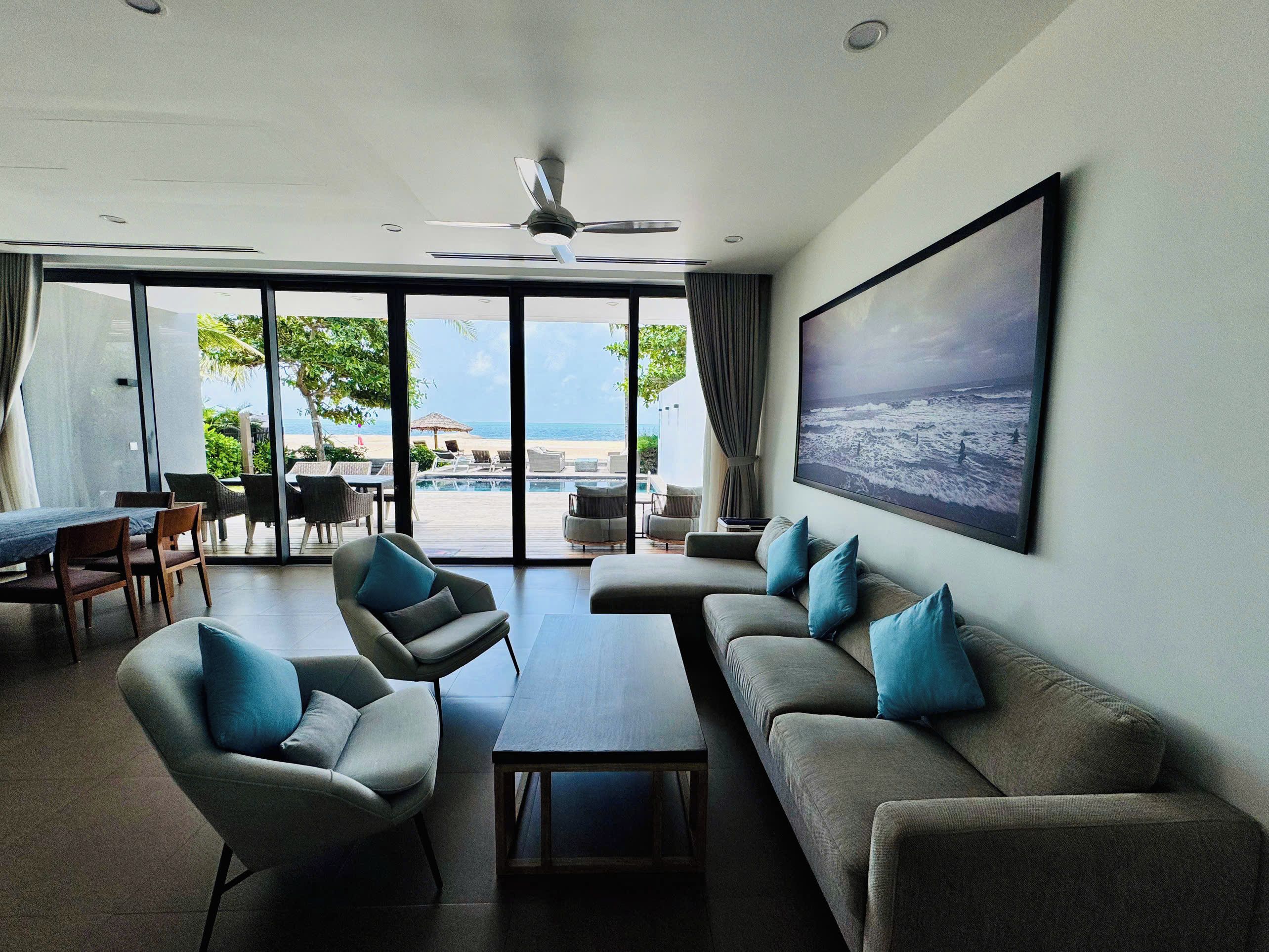 A modern living room with a large window, a ceiling fan, and a view of a pool and beach.